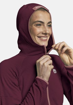 Frau lächelt, während sie eine dunkelrote Kapuzenjacke mit Logos auf der Kapuze und der Brust vor einfarbigem Hintergrund zumacht.