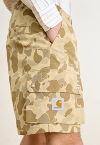Beżowe szorty w kamuflażu o gładkiej fakturze, z bocznymi kieszeniami i naszywką z logo Carhartt. Posiadają srebrne elementy do zapięcia kieszeni.