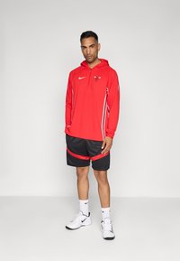 Rød Nike hettegenser med glidelås foran og logo, sammen med svarte shorts med røde detaljer. Hvite joggesko og sokker fullfører stilen.