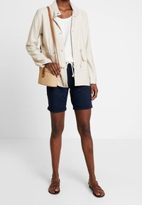 Světle béžová bunda s tkaničkou v pase, bílý top, námořnické šortky a hnědé sandály. Malá pletená crossbody taška přidává texturu.