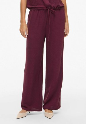 Pantalon large bordeaux en tissu texturé avec une taille à cordon réglable. Associé à des chaussures à bout pointu de couleur claire.