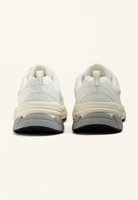 Bijele mrežaste tenisice s teksturiranom površinom, bež srednjim đonom i sivoj potplatu. Imaju povučnu traku na peti za lakše obuvanje.