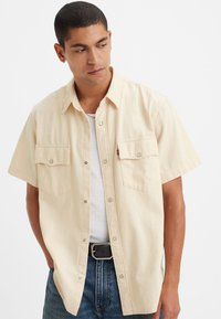 Short-sleeved beige button-up shirt made of cotton, featuring two chest pockets, a classic collar, and silver snap buttons.
