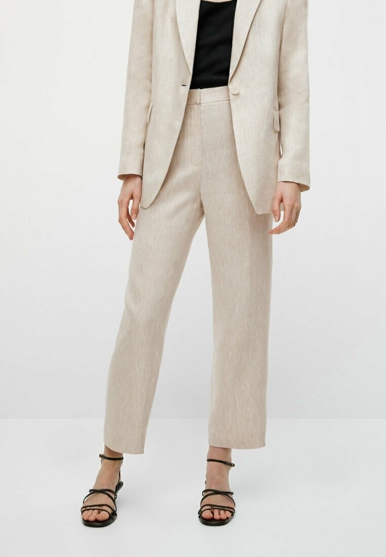 Costume en lin beige clair avec un blazer ajusté et un pantalon droit, associé à des sandales noires à lanières. Tissu texturé.