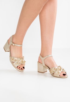 Sandales à talons carrés pailletés dorés avec embellissements perles et perles sur les brides croisées. Conception à bout ouvert avec une bride de cheville réglable.