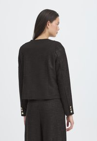 Giacca nera testurizzata con silhouette dritta, scollo rotondo e maniche lunghe. Presenta dettagli con bottoni dorati sui polsini.