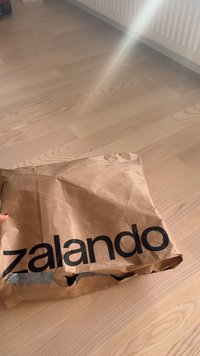 Gyűrött barna papírdoboz fekete "zalando" logóval, a tetején ragasztószalaggal, világos fa padlón pihen.