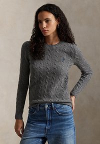 Mujer con cabello rizado que lleva un suéter gris de punto trenzado y vaqueros azules, de pie con una mano en la cadera frente a un fondo liso.