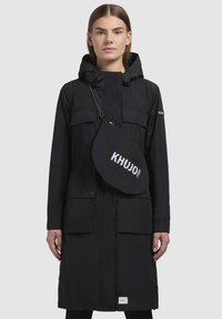 Parka neagră lungă cu glugă, având două buzunare frontale, închidere cu fermoar și o geantă mică tip crossbody cu inscripția "KHUJO 2010" imprimată în alb.