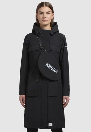 Schwarzer langer Parka mit Kapuze, ausgestattet mit zwei Fronttaschen, einem Reißverschluss und einer kleinen Umhängetasche mit dem Aufdruck "KHUJO 2010" in Weiß.