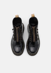 Paire de bottes en cuir noir brillant avec fermetures éclair à l'avant, coutures jaunes sur les semelles et languettes beiges sur fond blanc.