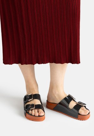 Füße in schwarzen Lackledersandalen mit zwei großen Schnallen und dicken braunen Sohlen, kombiniert mit einem roten gerippten Midirock.