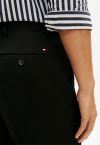 Οι μαύρες παντελόνες διαθέτουν πλαϊνή τσέπη με μαύρο κουμπί και ένα μικρό κόκκινο, λευκό και μπλε λογότυπο. Είναι ορατό ένα ριγέ μαύρο και λευκό πουκάμισο.