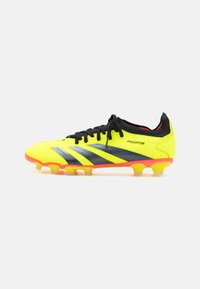 Botín de fútbol amarillos brillantes con rayas negras, parte superior sintética texturizada, cordones negros y suela con acentos naranjas y múltiples tacos.