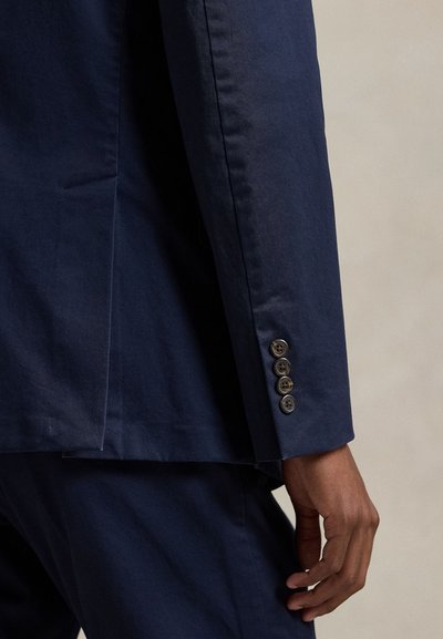 Detail osoby nosící tmavě modrý oblek s čtyřmi knoflíky na manžetě a odpovídající tmavě modré kalhoty.