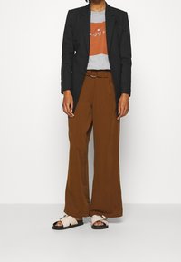 Persoon in een zwarte blazer, grijs en roestkleurig graphic t-shirt, bruine wide-leg broek met riem en crèmekleurige slidesandalen met zilveren gespen.