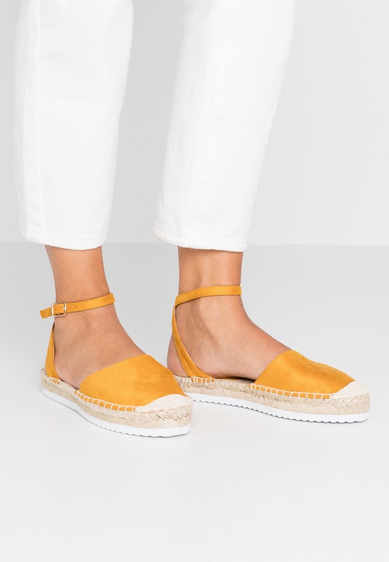 Pieds portant des sandales espadrilles jaune moutarde avec brides cheville et pantalon blanc sur fond uni.