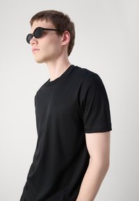 T-shirt nera a maniche corte, texture liscia, scollo rotondo. Accompagnata da occhiali da sole rotondi, scuri, con montatura sottile.