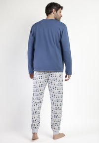 Hombre descalzo vistiendo una camiseta de manga larga azul y pantalones de pijama gris claro con patrones de personajes de dibujos animados en negro y naranja, de pie contra un fondo blanco.