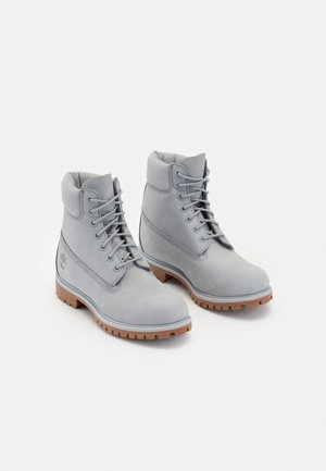 Paire de bottines basses en cuir gris clair à lacets, avec col rembourré et semelles crantées en caoutchouc marron sur fond blanc.