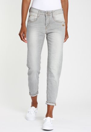 Femme portant un jean gris clair avec revers et des baskets blanches, debout devant un fond blanc uni.