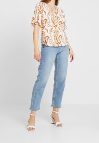 Haut blanc avec motif paisley orange, manches courtes volantées, associé à un jean droit bleu clair et des sandales à talons blocs blanches.