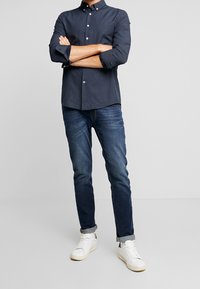 Man som står med armarna i kors, iklädd en mörkt mönstrad skjorta med knappar, rullade blå jeans och vita sneakers framför en enfärgad bakgrund.