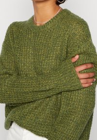 Grönstickad tröja med lös passform, strukturerad väv, långa ärmar och ribbade muddar. Lätt varierande nyanser av grönt förstärker dess djup.