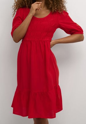 Femme aux cheveux bouclés portant une robe rouge arrivant aux genoux avec des manches bouffantes et un corsage smocké, posant avec une main sur la hanche et l'autre près du menton.