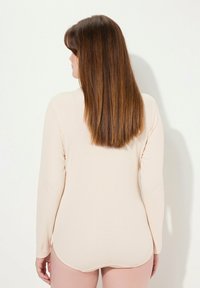 Langärmliger, figurbetonter Body aus hellbeigem Stoff. Glatte Textur mit abgerundetem Saum und subtilen Nähten entlang des Halsausschnitts und der Ärmel.