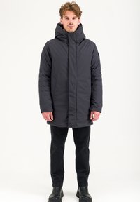 Giacca imbottita grigio scuro con colletto alto, zip frontale e tasche laterali. Indossata sopra pantaloni neri e stivali, caratterizzata da una texture liscia.