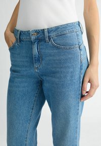Modri jeans s visokim pasom, petimi žepi in kontrastnim šivom, z bližnjim posnetkom pasu in žepov.