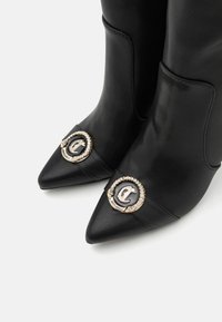Crne kožne čizme s uskim vrhom imaju zlatne i crne kružne detalje s logotipom, glatku teksturu i suptilne šavove.