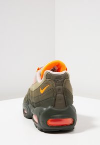 Boty Nike Air v olivově zelené a oranžové barvě, zobrazení zadní a boční části s viditelnou vzduchovou vložkou v podrážce na bílém pozadí.