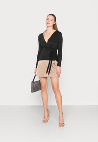 Top preto com mangas longas, decote em V profundo e detalhe de laço na cintura. Combinado com uma saia bege e uma mala preta texturizada.