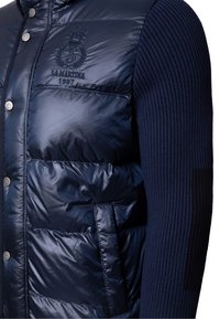 Smanicato imbottito blu navy con maniche in maglia a coste, realizzato in tessuto lucido, con bottoni a pressione argentati e logo ricamato sulla parte frontale.