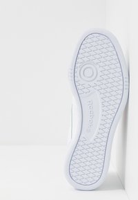 Vit Reebok-sneakersula med diamantmönster och cirkulär greppdetalj, placerad vertikalt mot en vit hörnvägg.