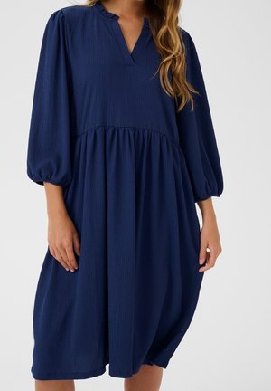 Kobieta nosząca luźną granatową sukienkę do kolan z rękawami do łokcia z bufkami i dekoltem w serek z małym kołnierzykiem.