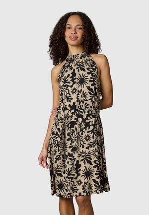 Frau mit lockigem Haar, die ein beiges ärmelloses Kleid mit schwarzem abstraktem Blumenmuster und hohem Ausschnitt trägt, steht vor einem schlichten Hintergrund.