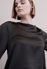 Top en satin noir avec une texture lisse, dotée d'un col bateau et de manches longues, porté par un modèle sur un fond neutre.