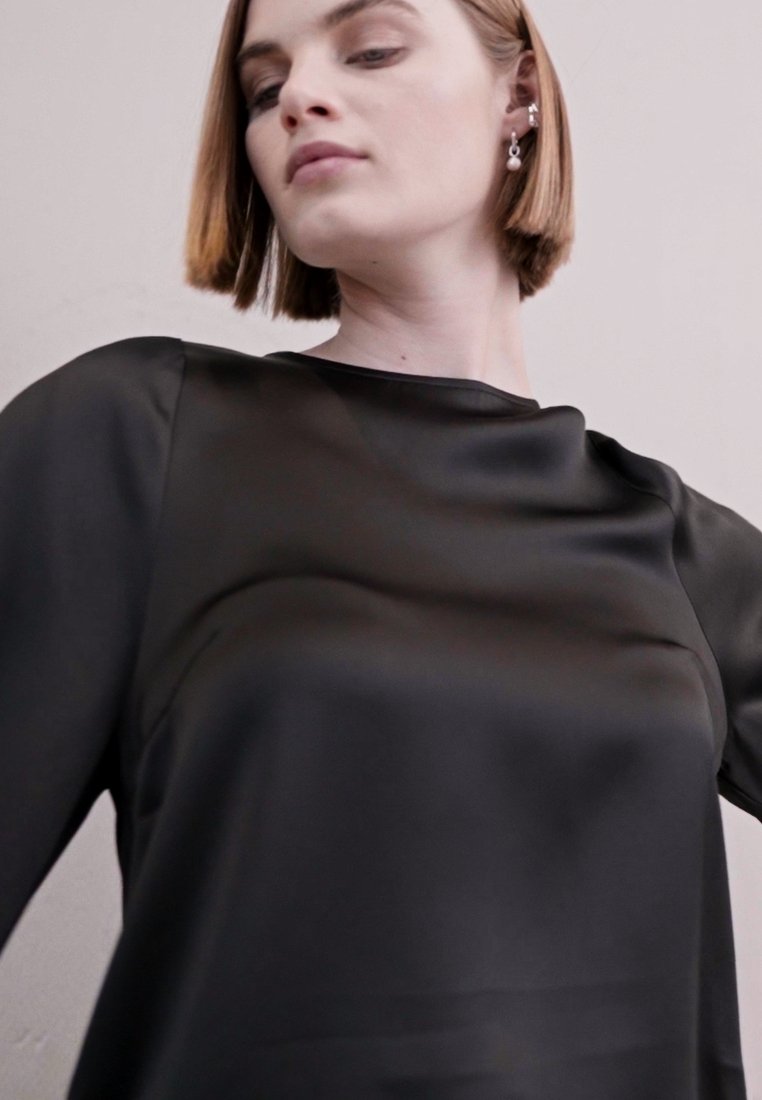 Top en satin noir avec une texture lisse, dotée d'un col bateau et de manches longues, porté par un modèle sur un fond neutre.