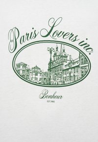 Λευκό T-shirt με πράσινη γραμμή που απεικονίζει πολυκατοικίες στο Παρίσι, με την επιγραφή «Paris Lovers inc.» από πάνω και «Bonheur» από κάτω σε κομψή γραμματοσειρά.