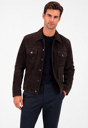 Hombre con chaqueta de ante marrón oscuro sobre una camisa negra y pantalones oscuros, de pie con una mano en el bolsillo contra un fondo blanco.