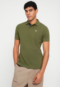 Zelená polokošile vyrobená z texturované látky, s límečkem, krátkými rukávy a logem na hrudi. Kombinovaná s béžovými šortkami.