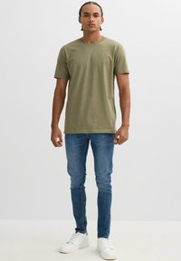 Camiseta de algodón verde oliva de manga corta y cuello redondo, combinada con jeans ajustados de mezclilla azul y zapatillas blancas.