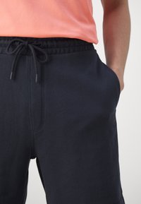 Tamsiai mėlyni medvilniniai sportiniai džinsai su elastingu juosmeniu ir raišteliu, su šoniniais kišenėmis ir maža logotipo etikete.