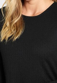 Svart topp med en strukturerad, ribbad tyg. Har rund halsringning och långa ärmar. Enkelt design utan synliga detaljer eller mönster.