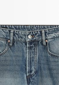 Jean en denim avec un lavage bleu délavé, doté d'une fermeture par bouton, cinq poches, des coutures visibles et une légère texture effilochée sur les bords.