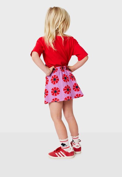 Rotes Kurzarmhemd kombiniert mit einem lila Rock, der mit roten Blumenmustern verziert ist. Rote Sneakers mit weißen Akzenten und gemusterte Socken.