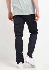 Man som bär svarta slim-fit-byxor, grå t-shirt och vita Adidas-sneakers, stående mot en enkel ljus bakgrund.